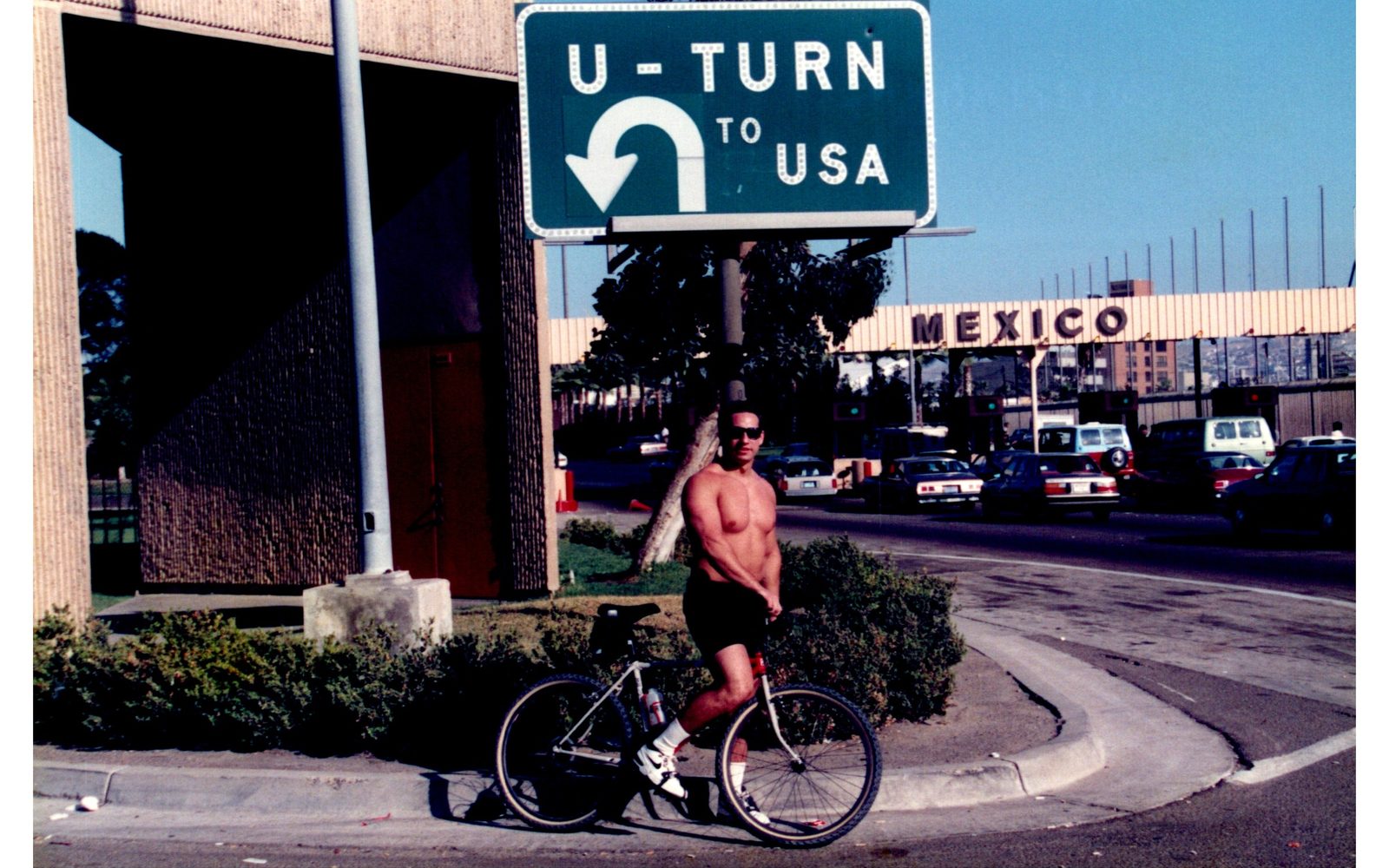 A young Ben standing with his bicycle beneath a road sign reading U-Turn to USA, with Mexico signage behind